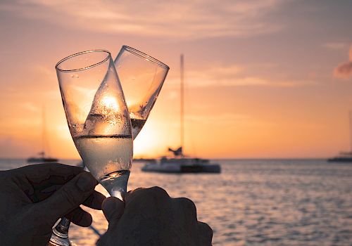 Two champagne glasses clink together against the backdrop of a serene sunset over a body of water with sailboats in the distance.