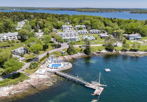 Aerial view of a waterfront resort with multiple buildings, a swimming pool, and a dock extending into a body of water surrounded by greenery.