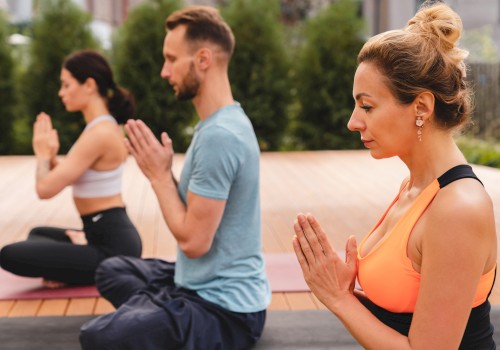 Three people are practicing yoga outdoors, sitting in a meditative pose with their hands in a prayer position, focusing on mindfulness and relaxation.
