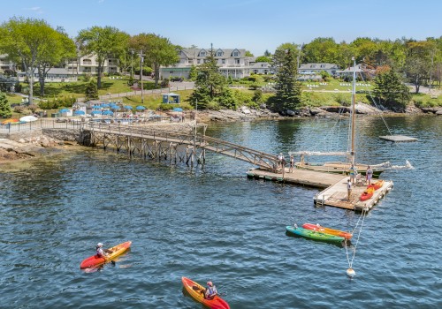 People kayaking near a wooden dock with sailboats, against a backdrop of houses and trees on the shore, under a clear blue sky.